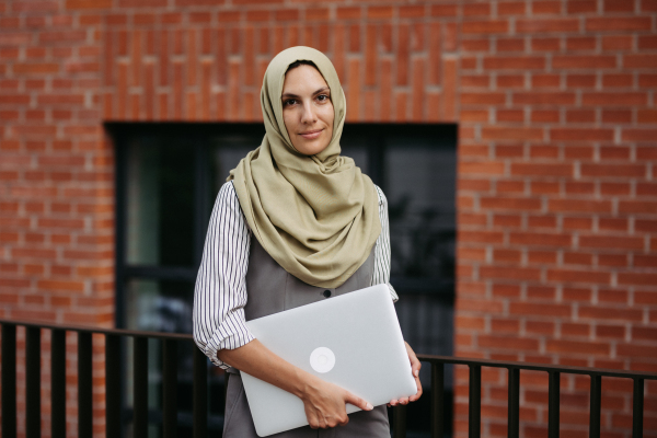 Portrait of beautiful woman in hijab standing on city street. Muslim businesswoman with laptop in hands. Iran, Afganistan female teacher in front of school building.