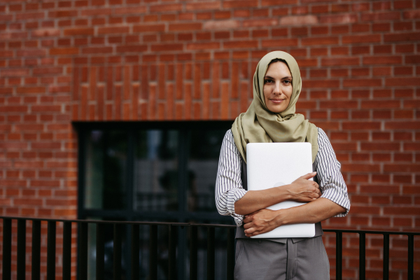 Portrait of beautiful woman in hijab standing on city street. Muslim businesswoman with laptop in hands. Iran, Afganistan female teacher in front of school building.