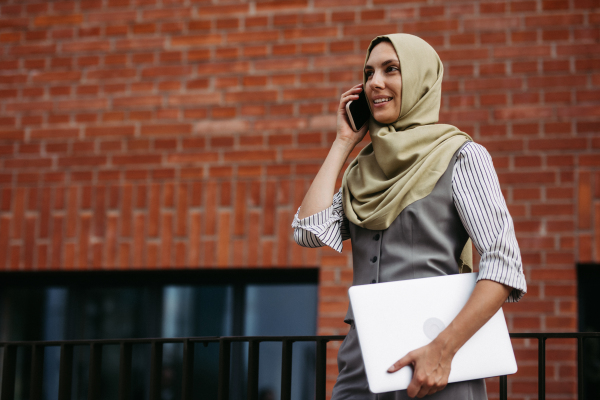 Portrait of beautiful woman in hijab standing on city street. Muslim businesswoman with laptop making call. Iran, Afganistan female teacher in front of school building.