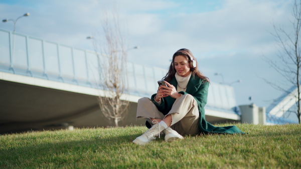 A beautiful young woman listening music, sitting in grass. Fashionable generation z woman with closed eyes enjoying cold spring day.