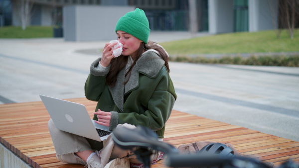 A sick university student on bench in front of university building blowing nose. Fashionable Gen z woman with allergy going to school.