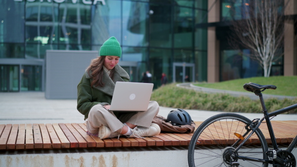 An university student on bench in front of university building working on laptop. Fashionable Gen z woman traveling to school by bike. City commuter student, sustainable lifestyle.