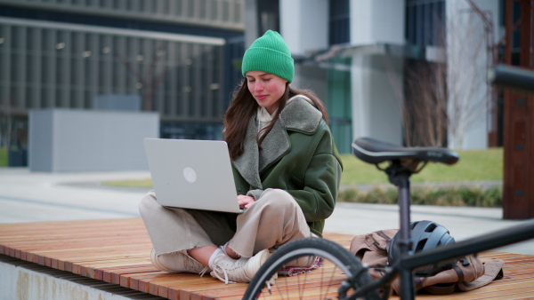 An university student on bench in front of university building working on laptop. Fashionable Gen z woman traveling to school by bike. City commuter student, sustainable lifestyle.