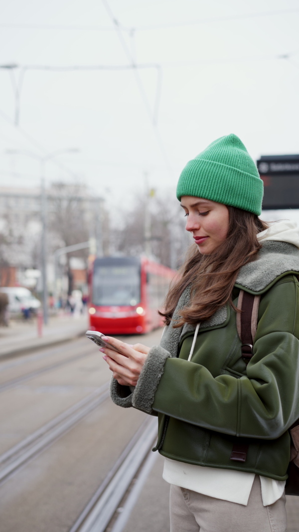 A beautiful university student waiting on bus stop, going to school. Fashionable Gen z woman standing on city street, scrolling on smartphone.