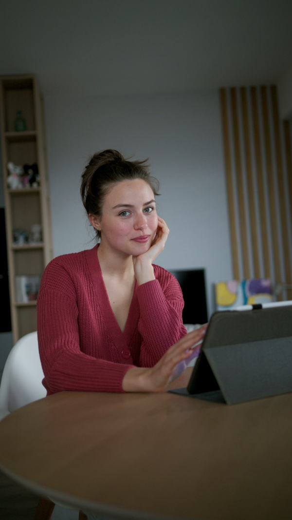 A woman sitting at table with tablet in front of her, thinking. Woman managing household functions of smart home system, lighting, security cameras, door locks, thermostat or heating settings.