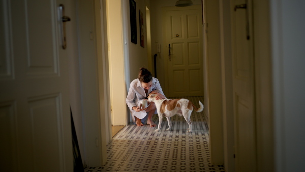 Woman in pajamas, playing with her cute dog, drinking cup of morning coffee. Weekend morning relaxation at home. Hygge lifestyle.