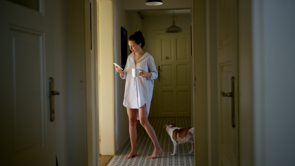 Young woman in pajamas in morning, carrying cup of coffee and returning back to bed. Side view of beautiful single woman at home alone over the weekend.