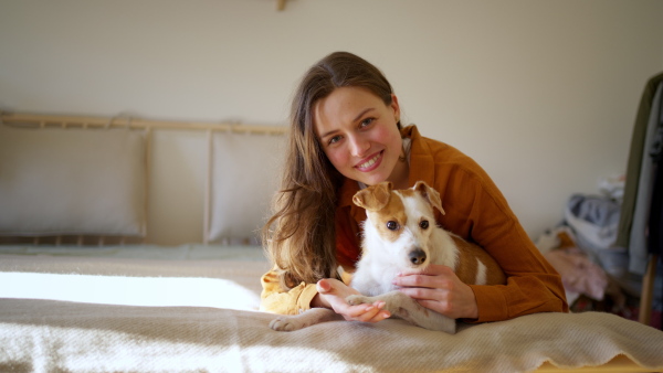 Young woman living alone in apartment. Beautiful single woman lying on bed, petting her dog, enjoying weekend. Looking at camera, smiling.