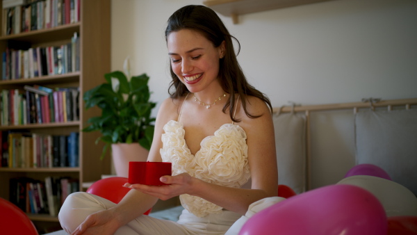 Beautiful woman sitting on bed in the middle of baloons, opening small present. Concept of Valentine's Day, love and romantic dating.