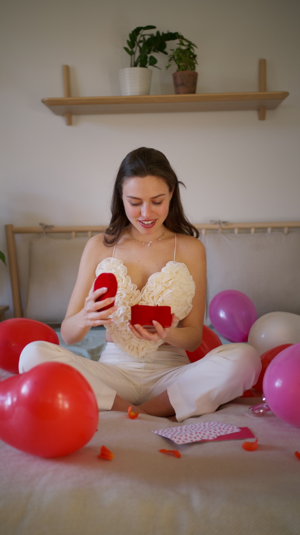 Beautiful woman sitting on bed in the middle of baloons, opening small present. Concept of Valentine's Day, love and romantic dating.