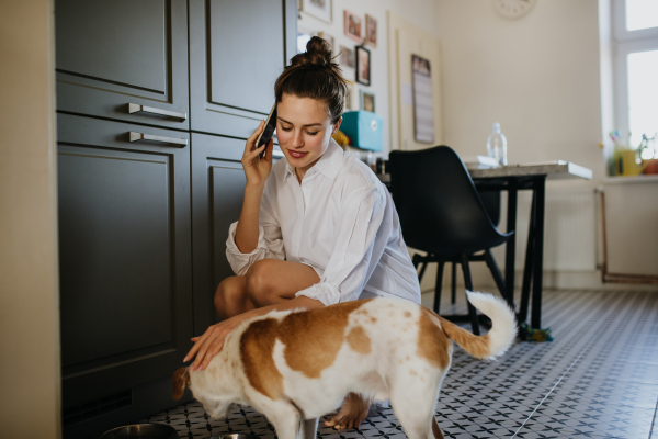 Woman in pajamas feeding dog and phone calling, smiling. Weekend morning routine at home. Hygge lifestyle for single woman.