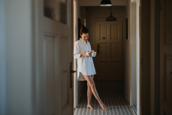 Young woman in pajamas in morning, carrying cup of coffee and returning back to bed. Side view of beautiful single woman at home alone over the weekend.