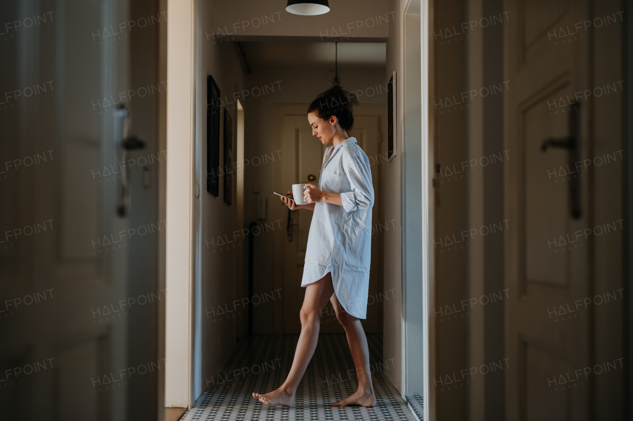 Young woman in pajamas in morning, carrying cup of coffee and returning back to bed. Side view of beautiful single woman at home alone over the weekend.