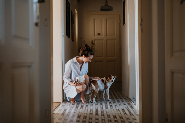 Woman in pajamas, playing with her cute dog, drinking cup of morning coffee. Weekend morning relaxation at home. Hygge lifestyle.