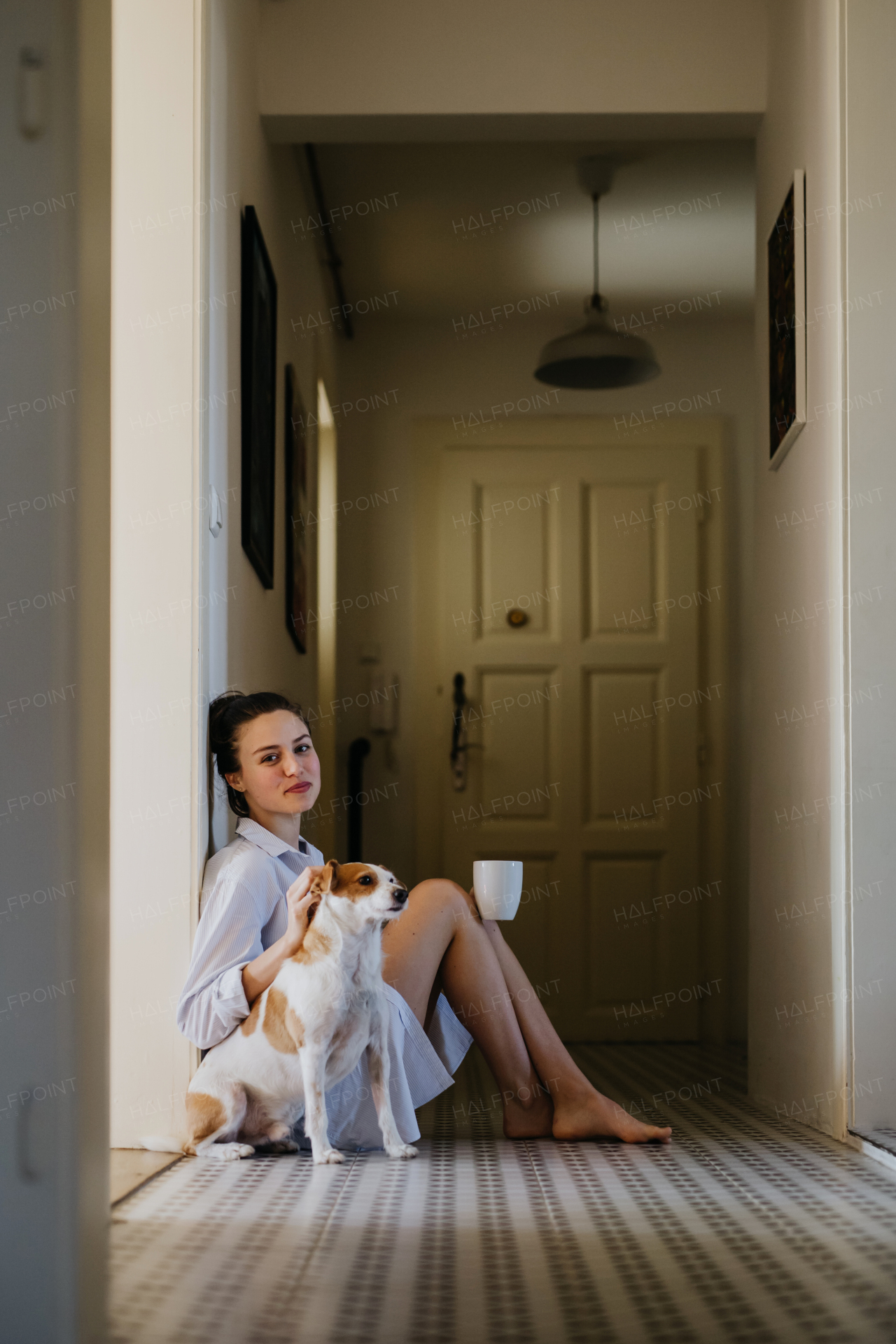 Woman in pajamas sitting on floor, playing with her cute dog, drinking cup of morning coffee. Weekend morning relaxation at home. Hygge lifestyle.