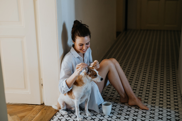 Woman in pajamas sitting on floor, playing with her cute dog, drinking cup of morning coffee. Weekend morning relaxation at home. Hygge lifestyle.