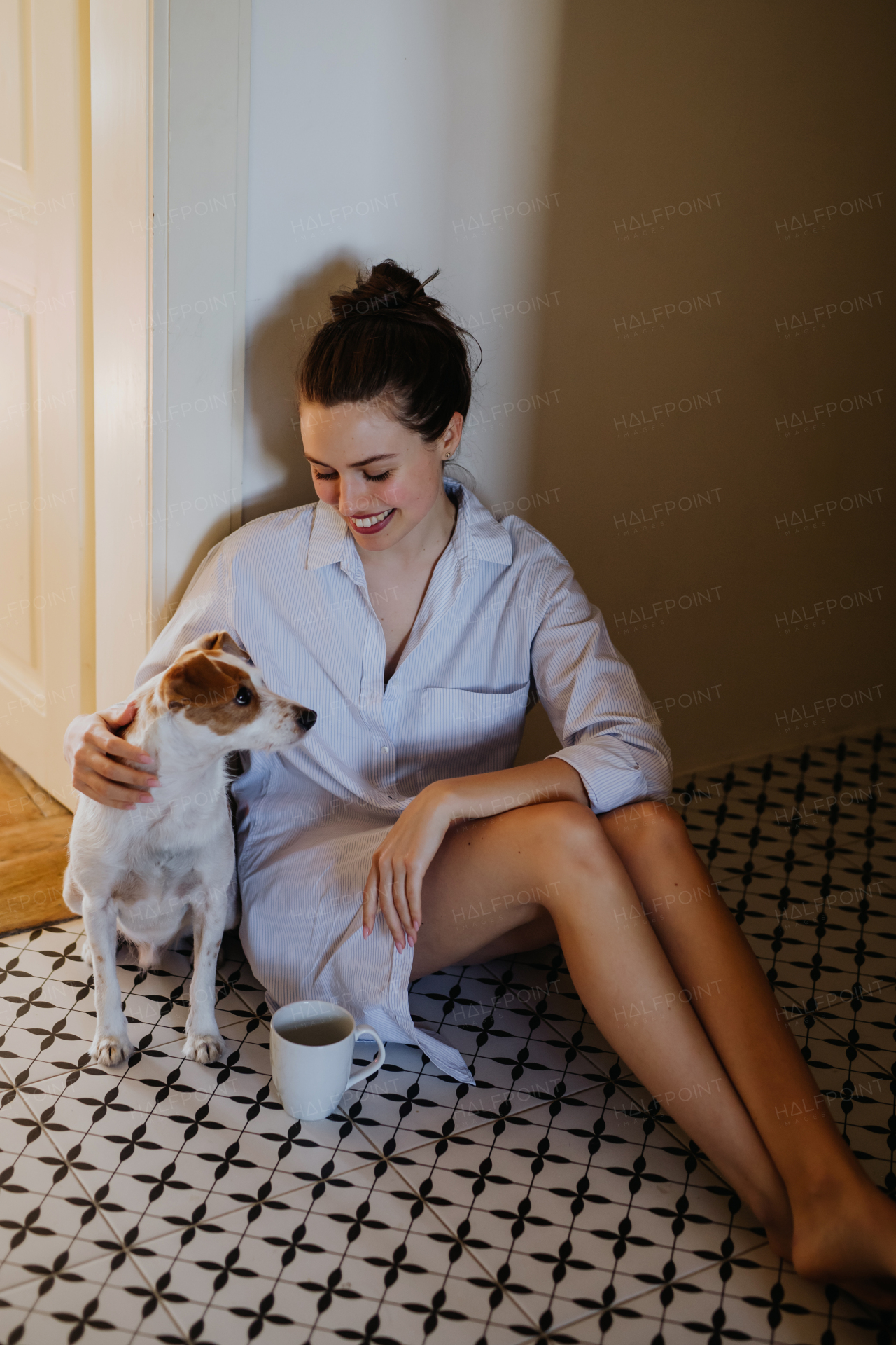 Woman in pajamas sitting on floor, playing with her cute dog, drinking cup of morning coffee. Weekend morning relaxation at home. Hygge lifestyle.