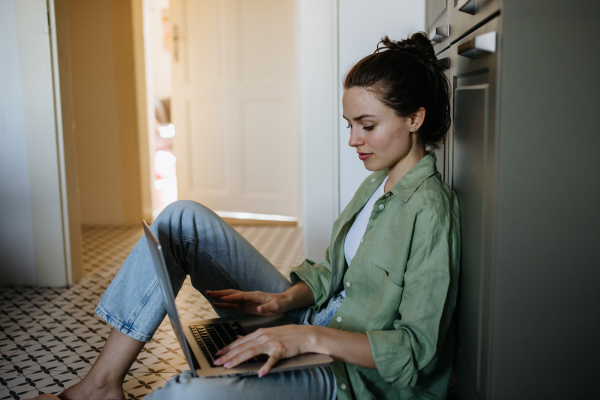 Young student e-learning on laptop at home. Businesswoman working remotely, sitting at kitchen floor and typing. Homeoffice for single woman living alone.