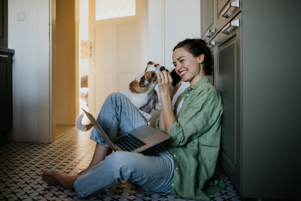 Young student e-learning on laptop at home, cute small dog wants to play. Businesswoman working remotely, sitting at kitchen floor, phone calling. Home office for single woman living alone.