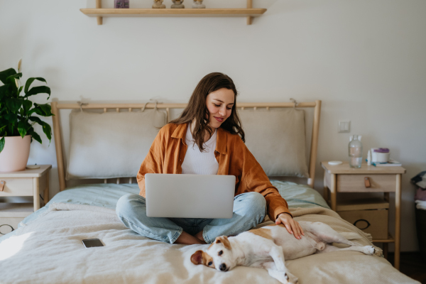 Young student e-learning on laptop at home, cute small dog besides. Businesswoman working remotely, sitting on bed. Home office for single woman living alone.