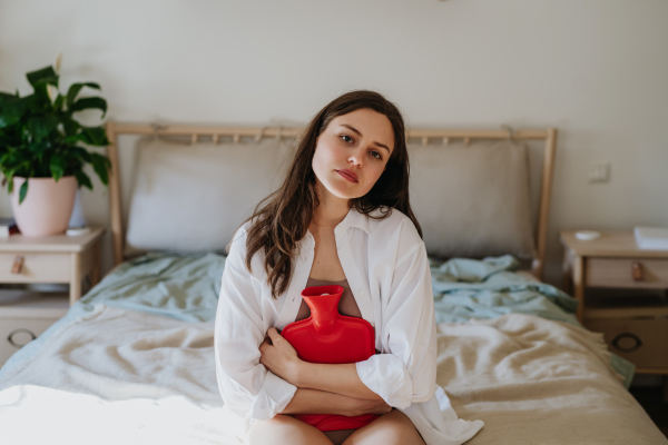 Young woman at home suffering from menstrual pain, having cramps. Woman warming lower abdomen with a hot water bottle, endometriosis, and conditions causing pain in tummy.