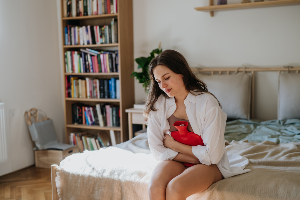 Young woman at home suffering from menstrual pain, having cramps. Woman warming lower abdomen with a hot water bottle, endometriosis, and conditions causing pain in tummy.