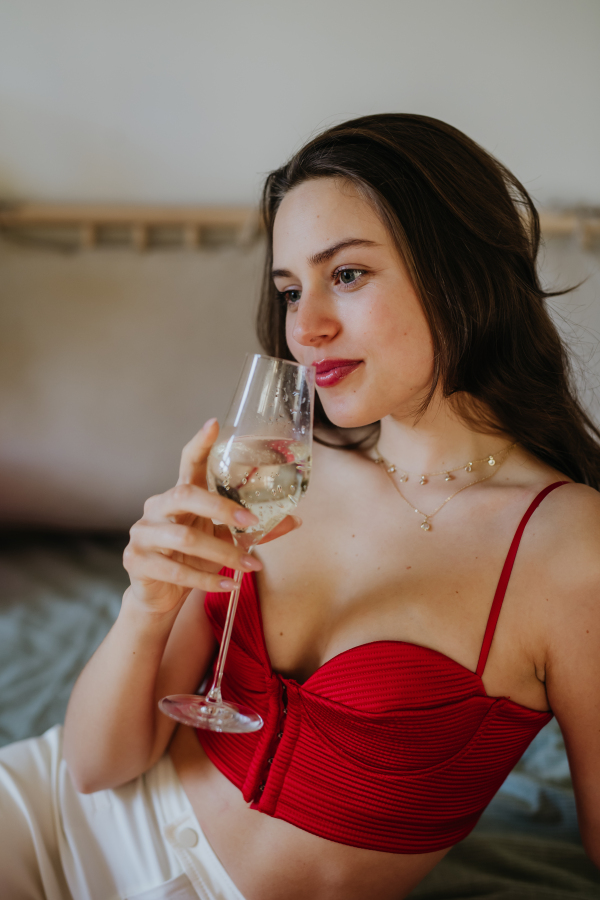 Romantic woman drinking champagne from a glass, dressed in a red attractive corset. Concept of Valentine's Day, love and romantic relationship.