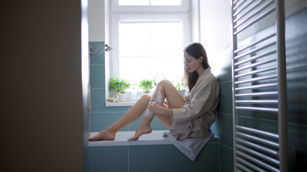 Beautiful woman applying moisturizing body lotion on her skin, legs. Skincare, beauty routine. Woman sitting in the bathroom on the bathtub, dressed in a bathrobe.