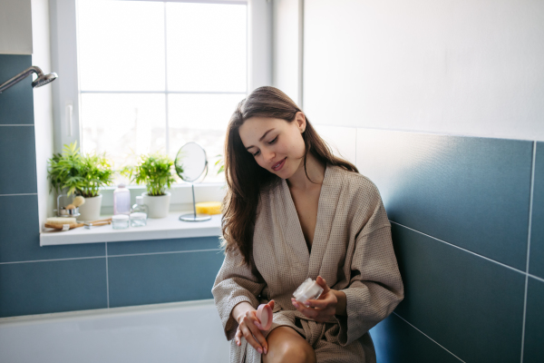 Beautiful woman applying moisturizing body lotion on her skin, legs. Skincare, beauty routine. Woman sitting in the bathroom on the bathtub, dressed in a bathrobe.