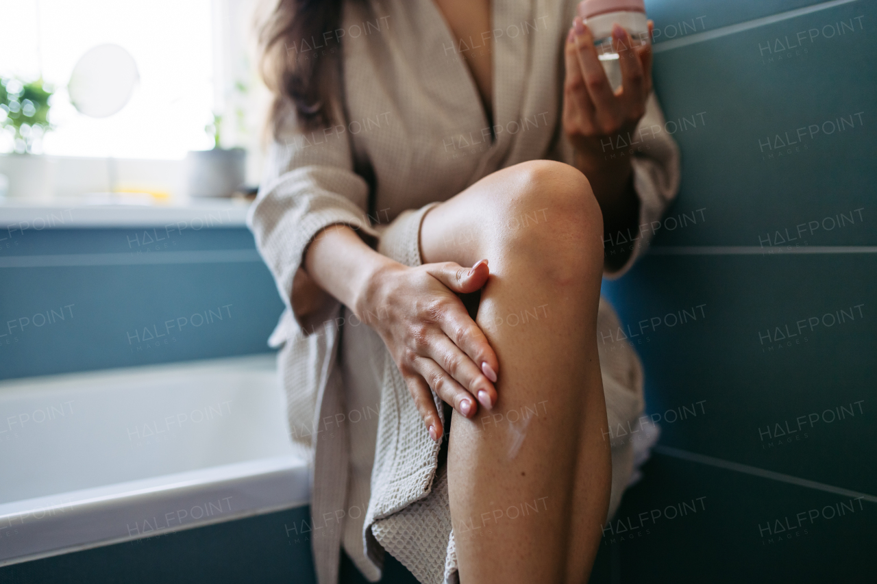 Beautiful woman applying moisturizing body lotion on her skin, legs. Skincare, beauty routine. Woman sitting in the bathroom on the bathtub, dressed in a bathrobe. Close up on leg.