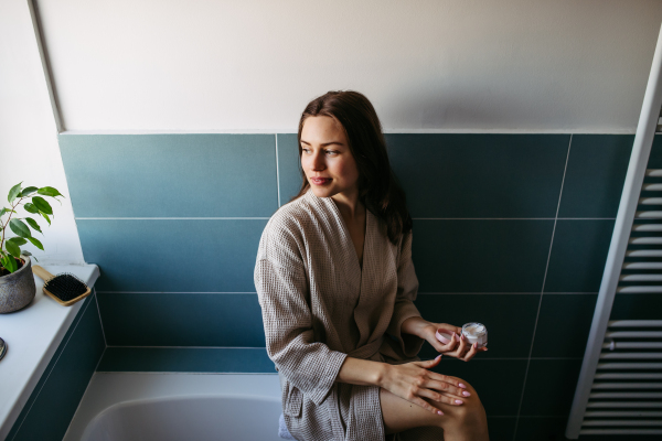 Beautiful woman applying moisturizing body lotion on her skin, legs. Skincare, beauty routine. Woman sitting in the bathroom on the bathtub, dressed in a bathrobe.