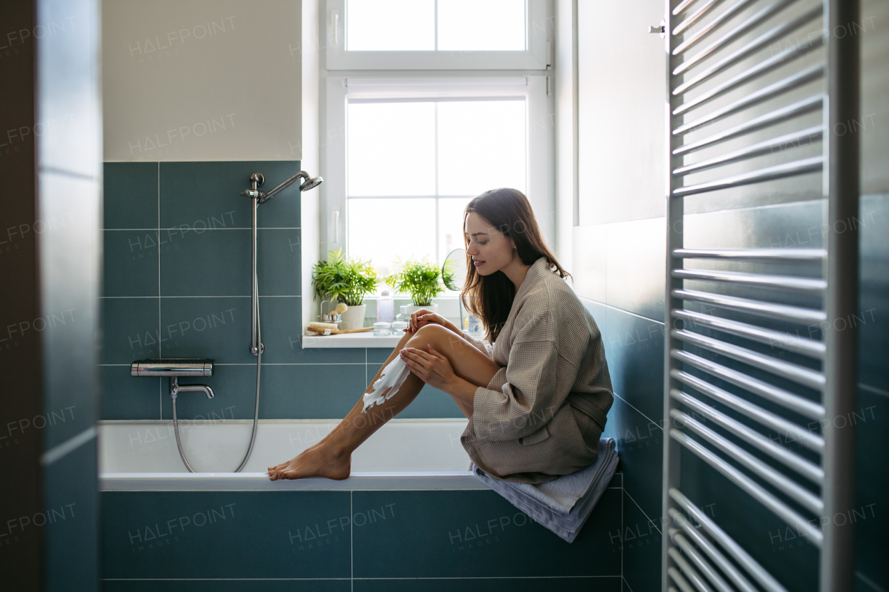 Side view of beautiful woman applying shaving foam, cream to legs, before shaving. Woman sitting on bathtube in bathroom, dressed in gray bathrobe.