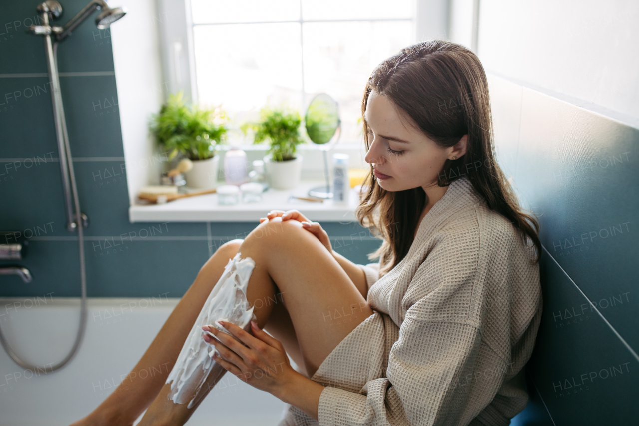 Side view of beautiful woman applying shaving foam, cream to legs, before shaving. Woman sitting on bathtube in bathroom, dressed in gray bathrobe.