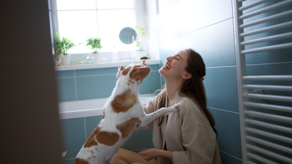 Side view of beautiful woman applying shaving foam, cream to legs, before shaving. Woman sitting on bathtube in bathroom, dressed in gray bathrobe.