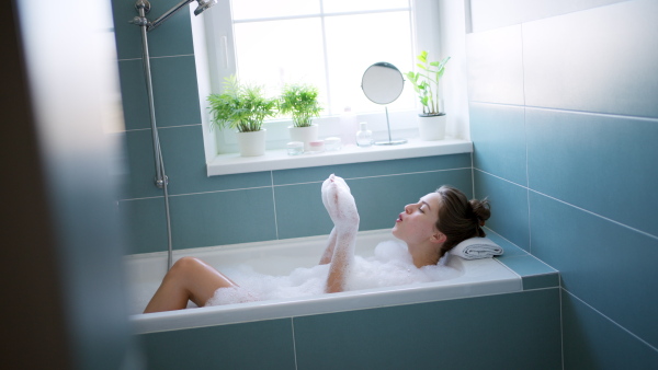 Beautiful woman shaving her legs with razor. Woman sitting on bathtube in bathroom, dressed in a bathrobe. Daily body and skin care routine.