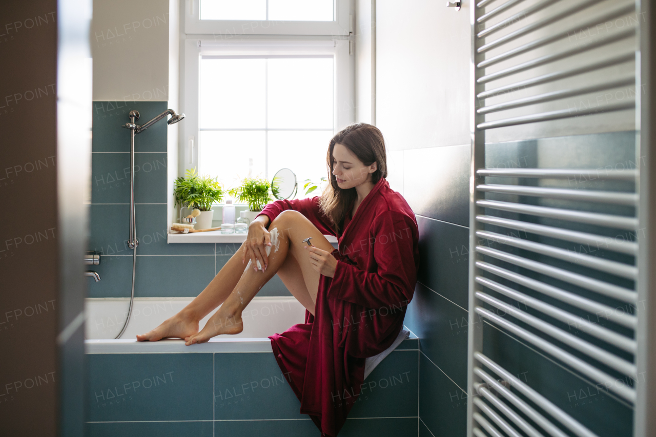 Side view of woman shaving her legs with razor, sitting on bathtube in bathroom. Wearing red bathrobe. Daily body and skin care routine.