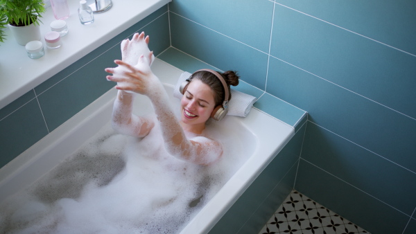 Woman shaving her legs with razor, sitting on bathtube in bathroom, looking at camera, smiling. Wearing burgundy bathrobe. Daily body and skin care routine.