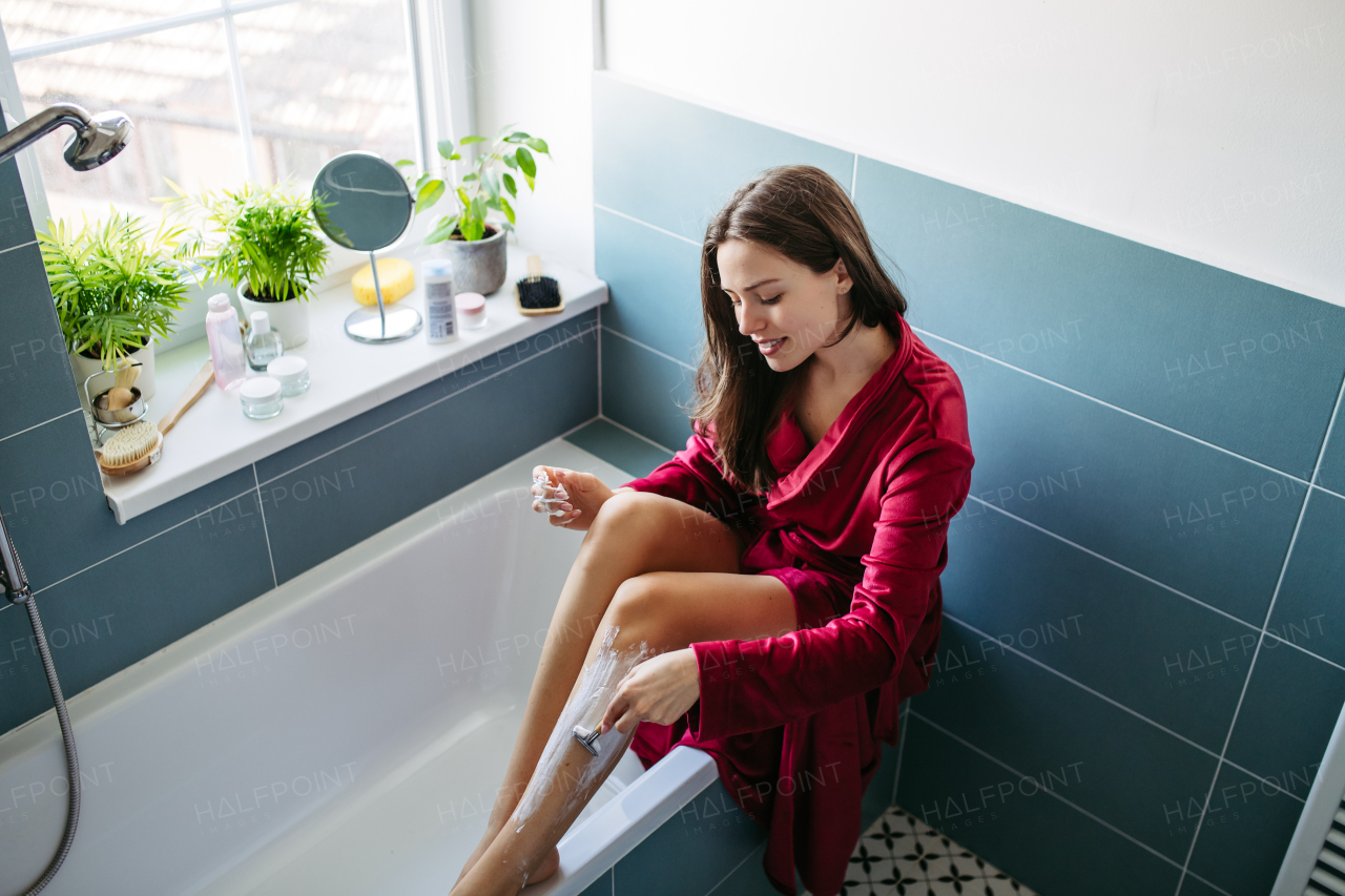 Woman shaving her legs with razor, sitting on bathtube in bathroom. Wearing red bathrobe. Daily body and skin care routine. High angle view.