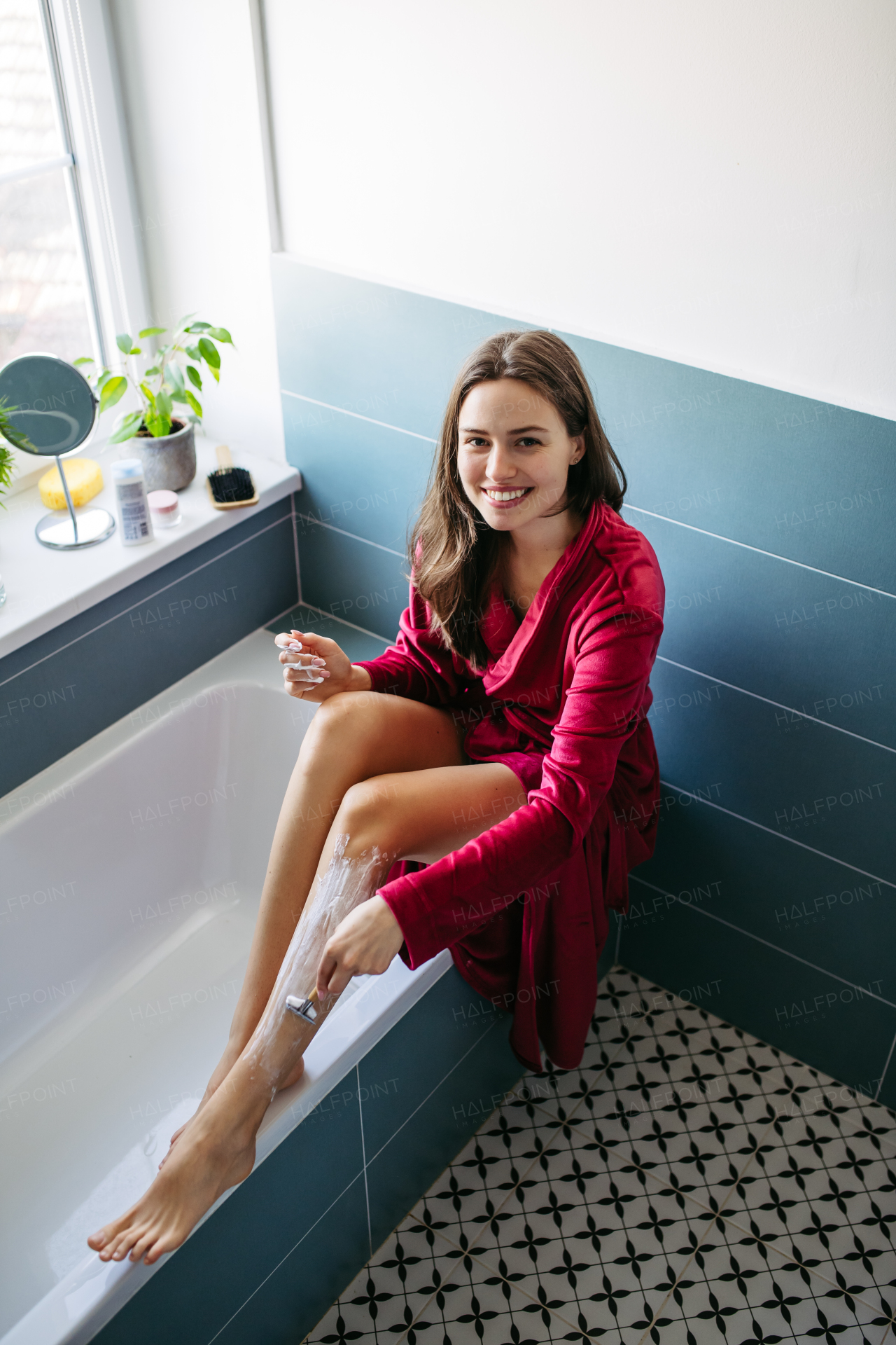 Woman shaving her legs with razor, sitting on bathtube in bathroom. Wearing red bathrobe. Daily body and skin care routine. High angle view.