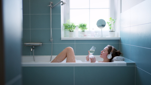 Woman shaving her legs with razor, sitting on bathtube in bathroom. Wearing red bathrobe. Daily body and skin care routine. High angle view.