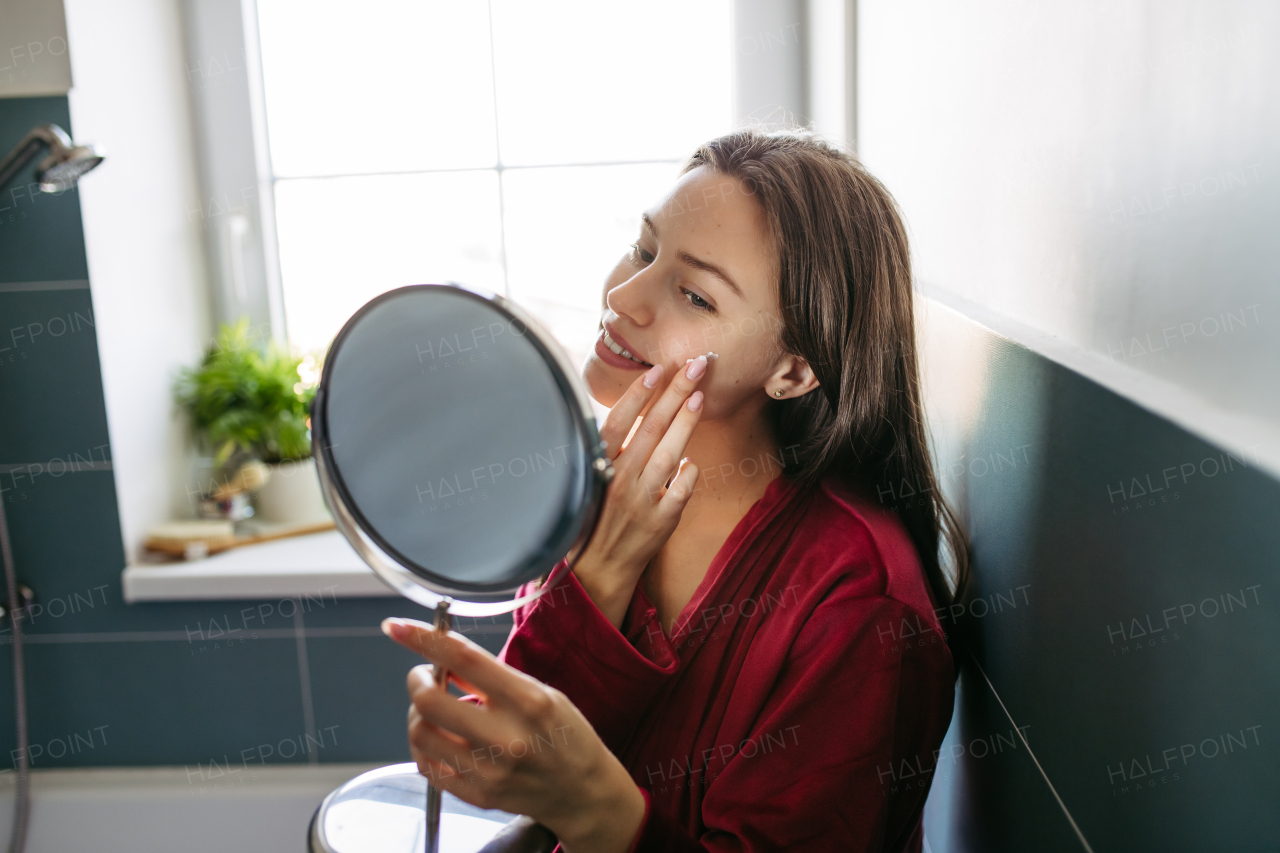 Beautiful woman applying facial moisturizer cream on face, looking into a vanity mirror, dressed in a burgundy bathrobe. Morning skincare routine.