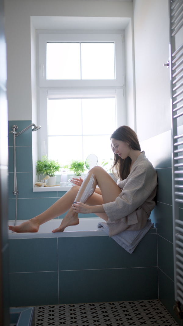 Beautiful woman applying facial moisturizer cream on face, looking into a vanity mirror, dressed in a burgundy bathrobe. Morning skincare routine.