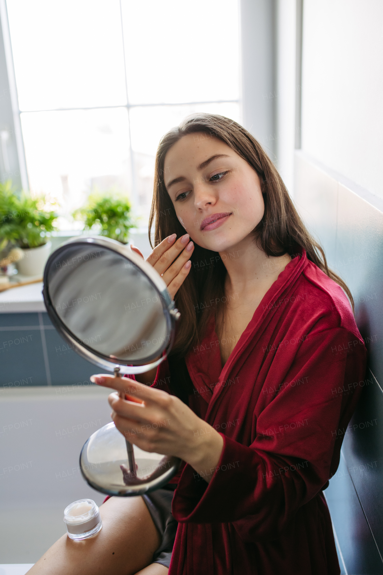 Beautiful woman applying facial moisturizer cream on face, looking into a vanity mirror, dressed in a burgundy bathrobe. Morning skincare routine.