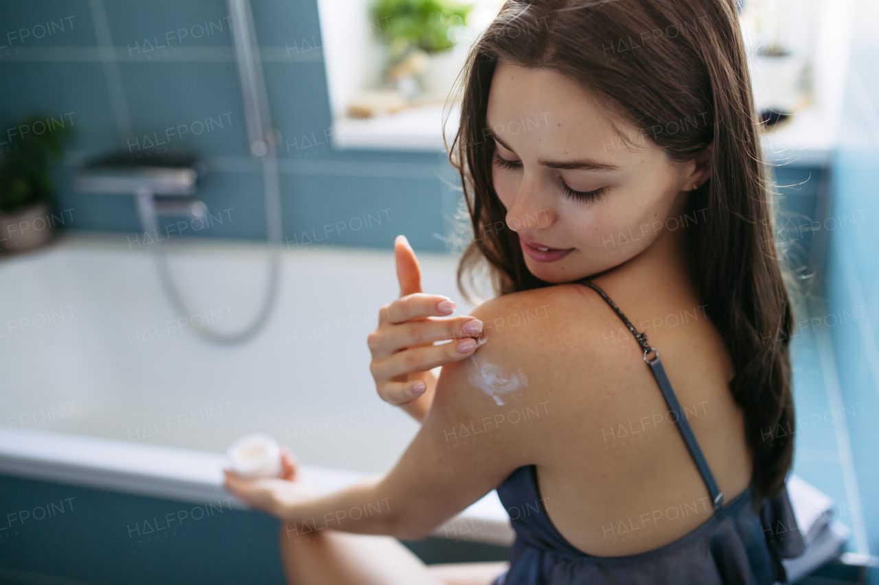 Young beautiful woman applying moisturizing body lotion to shoulder after showering. Skincare, body care routine.