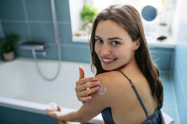 Young beautiful woman applying moisturizing body lotion to shoulder after showering. Skincare, body care routine.