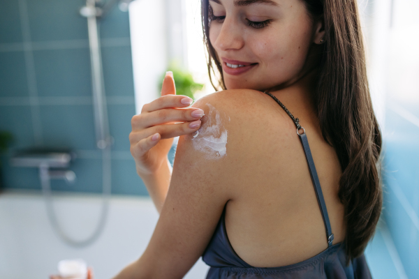 Young beautiful woman applying moisturizing body lotion to shoulder after showering. Skincare, body care routine.