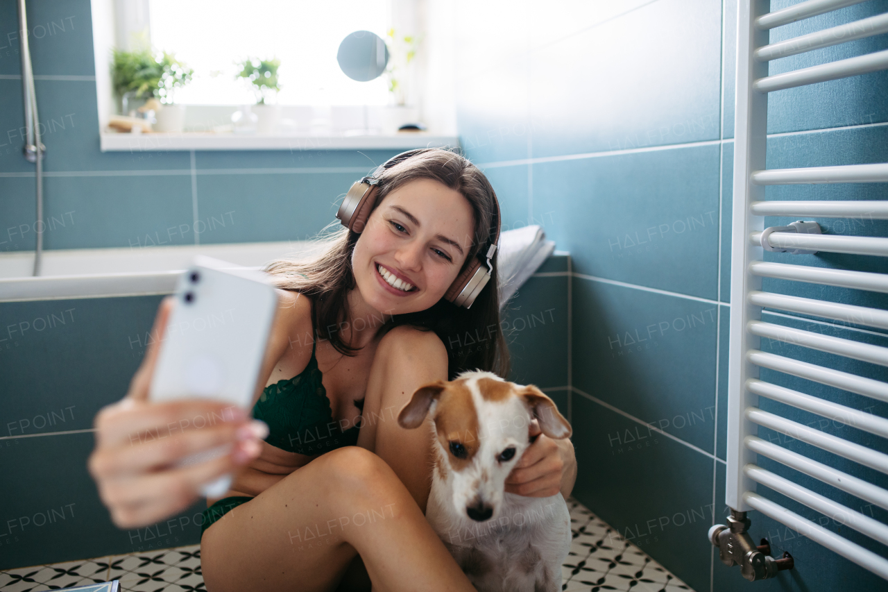 Young woman playing with a dog in the bathroom, getting ready for a bath in the tub. Relaxing and listening music after long work day.