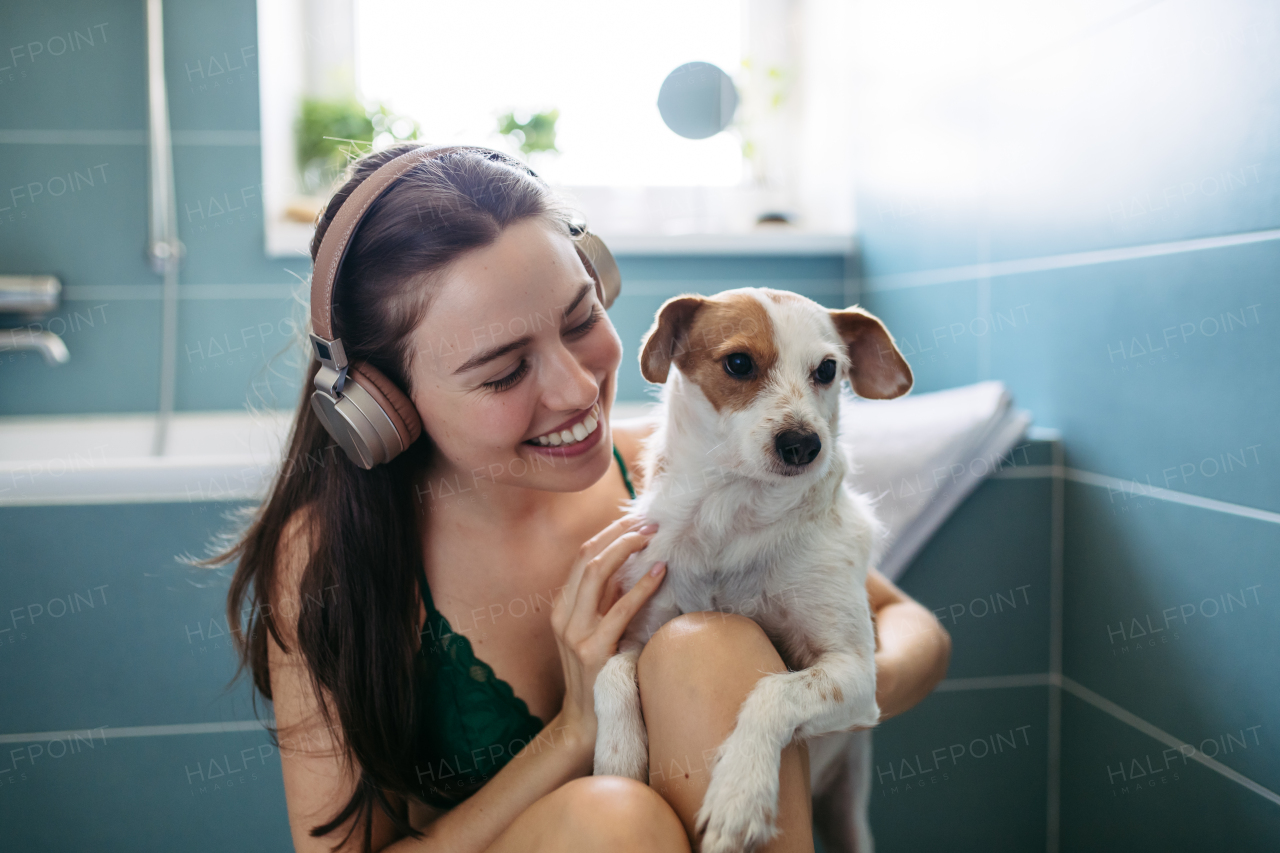 Young woman playing with a dog in the bathroom, getting ready for a bath in the tub. Relaxing and listening music after long work day.