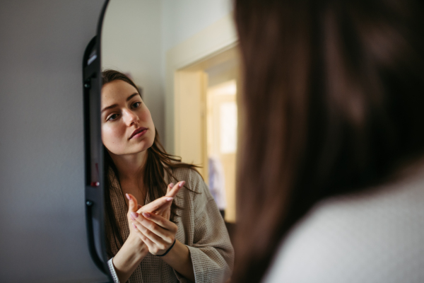 Beautiful young woman looking into mirror, applying moisturizer cream. Morning skincare routine.