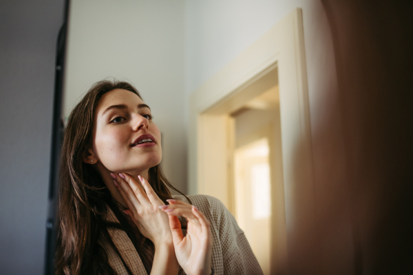 Beautiful young woman looking into mirror, applying moisturizer cream on face. Morning skincare routine.